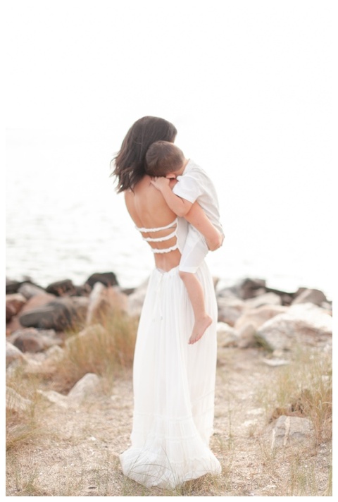 Mom and son look at the ocean as they embrace. Mom wearing long white gown with open back. 