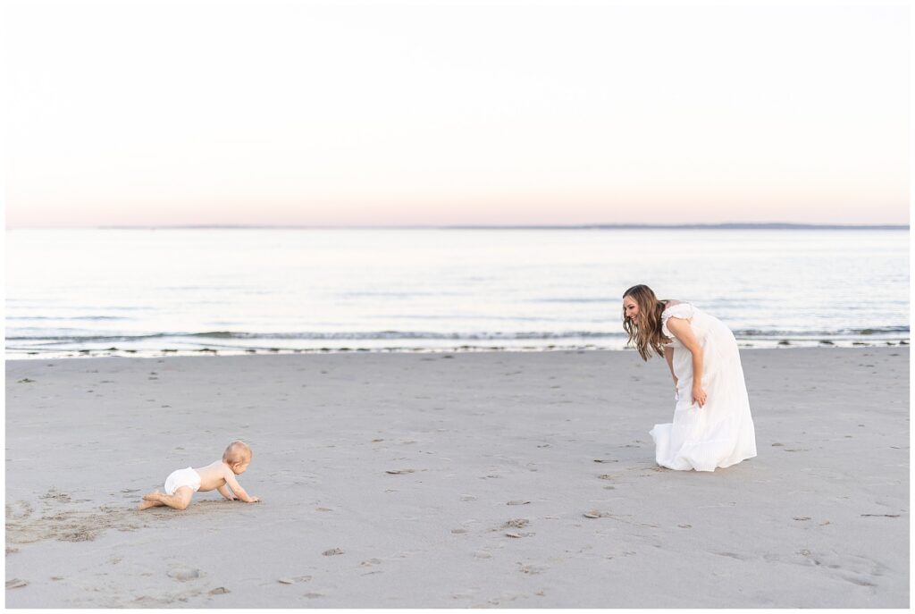 Gorgeous mom in white dress playing with her baby on the beach at Tod's Point in Greenwich. Baby crawling in the sand to his mama as they laugh and play during family portraits on the CT coast. 