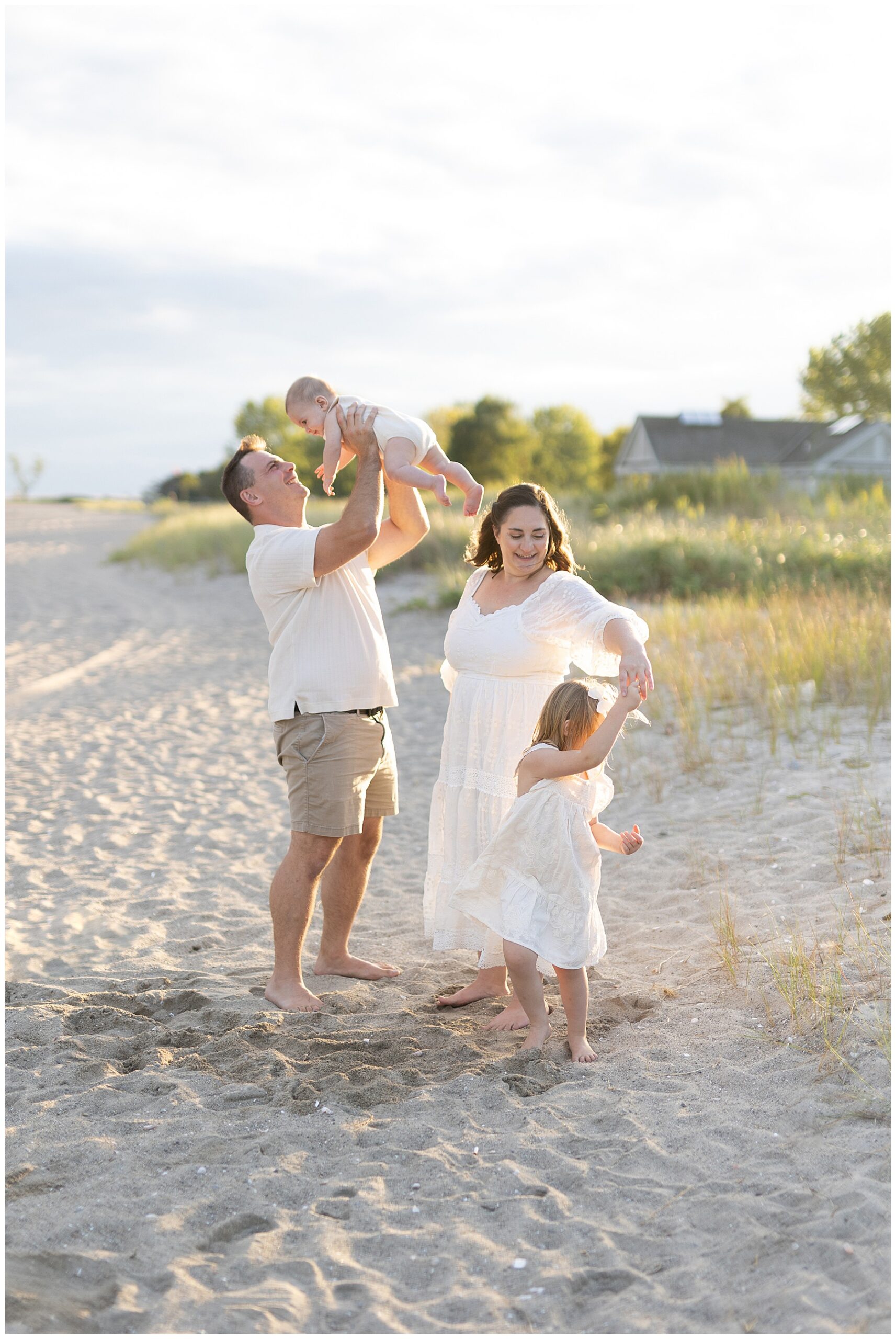Joyful family playing on the beach during their Westport beach portraits. Dad holding 6 month son in the air and mom twirling 3 year old daughter. Playful family portraits on the Connecticut Coast.