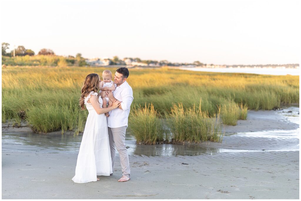 Variety a scenery for family beach photography session in Greenwich CT. Mom, dad and baby wear neutral tones, white, linen and gray chinos for family photos on the beach in CT.